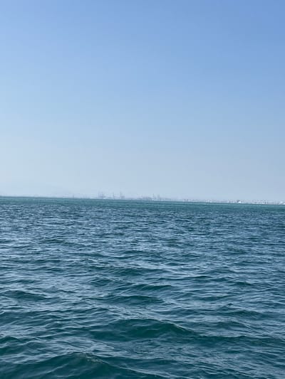 The image shows a wide view of the sea with a distant coastline. The water in the foreground is a deep blue-green, with visible ripples and waves. In the distance, a hazy coastline is visible, with a skyline of industrial structures, including what appear to be cranes, suggesting a port or industrial area. There are also faint outlines of buildings and possibly mountains or hills further inland. The sky above is a pale blue, with no clouds. The overall impression is one of a vast expanse of water with a busy, industrialized area in the background. This scene is consistent with the port of Callao, Peru, which is known for its extensive shipping facilities and industrial activity.