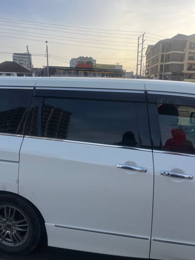 The image captures a street scene in Gitothua ward, Kenya, featuring a white vehicle on the right and an urban landscape in the background. The vehicle, likely a minivan, shows part of its side body, chrome door handles, dark tinted windows, and a multi-spoke wheel. Reflections are noticeable in the vehicle's windows; a person wearing a red top is faintly visible inside, reflected in the rear window, while a more indistinct figure is seen in the side window. A building is also visibly reflected in the tinted glass.
The background reveals a street lined with buildings. Overhead, numerous power lines crisscross the sky, supported by utility poles. The sky appears hazy with a yellowish hue, suggesting either early morning or late afternoon. On a building in the mid-ground, bright signs are visible, displaying the text "DJ HO LOUNGE" and "HOURI," indicating a commercial establishment, possibly a bar or entertainment venue. The overall setting is an urban or semi-urban environment.