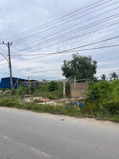 The image captures a roadside scene in Bundar, Indonesia. The foreground features an asphalt road with a white painted line. Beyond the road, a patch of overgrown grass and vegetation borders an area with remnants of a makeshift structure. This structure appears to be a partially dismantled stall or shelter, with wooden posts and a blue tarp visible. Scattered debris, including what looks like a mattress or large cushion, blue plastic sheeting, and other refuse, lies on the ground within the vicinity of the structure. In the background, a large tree stands prominently, with more greenery and palm trees visible further in the distance. Utility poles and overhead power lines traverse the sky, which is overcast with gray clouds. The overall impression is one of a rural or semi-urban environment, possibly undergoing development or in a state of disrepair.