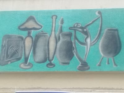 A horizontal mural is painted directly onto a textured wall in Montevideo, Uruguay. The artwork features seven stylized objects rendered in shades of grey and beige against a uniform teal background. From left to right, the depicted objects are: a rectangular electronic device, resembling a tablet or e-reader, displaying a stylized hand or abstract pattern on its screen; a mushroom-shaped lamp or decorative object with a slender stem and flared base; a lidded, stout cylindrical jar; a tall, slender bottle with a narrow neck and small foot; a short, bulbous vase; an abstract, metallic-appearing sculpture characterized by a central body with multiple curving, elongated appendages, topped with a sphere; and a rounded, three-legged pot. The mural occupies a distinct band on the wall, bordered above by a rough white wall section and below by a flat, light beige horizontal architectural element, consistent with a window sill or ledge. There are no people, actions, or direct interactions depicted within the scene.