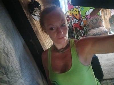This image depicts a single adult female, approximately 30-40 years of age, located in an indoor or sheltered environment within Santa Fe, United States. The female, positioned in the foreground and center, has light skin and reddish-blonde hair pulled back. She is wearing a lime green tank top and a multi-strand, silver-colored necklace. Her right arm is extended, indicating she is holding the camera to take a self-portrait. Her head is tilted slightly to her right, and her eyes are directed toward the upper left of the frame.
The immediate background consists of multiple surfaces. To the left of the female, a light-colored fabric with vertical stripes, resembling a sheet or blanket, is draped or hanging. Behind her, a wall constructed of horizontal wooden planks is visible. A black and white baseball cap with a tag attached is hanging on this wooden surface. To the female's right, a brightly colored fabric or poster featuring a pop-art design, including the word "BOOM!", stars, and stylized city buildings, is displayed. Further to the right and slightly behind the female, another patterned fabric with a muted, possibly geometric or abstract design is present. A dark, indistinct object, potentially a limb or part of the camera apparatus, partially obscures the top right corner of the frame. The lower right portion of the image suggests an unpaved ground surface.