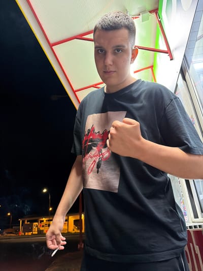 The image shows a young male standing outdoors at night, holding a lit cigarette in his left hand and forming a fist with his right hand. He is wearing a black t-shirt with a graphic design on it. The background suggests an urban or roadside setting with a building illuminated in the distance and streetlights visible. The overall lighting is dim, indicating it is nighttime. The location in the city of Giurgiu, Romania, is not discernible from the visual information provided in the image.
