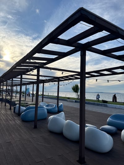 This media file captures a modern waterfront promenade in Taman, Russia, during the late afternoon or early evening. The sun is low in the sky, casting a soft, warm light from the bottom left of the frame and creating a gentle backlight effect against the sky and clouds.

The primary subject is a dark-stained wooden pergola, which runs horizontally across the upper and middle portions of the scene, creating a sense of depth and perspective. The pergola has an open lattice roof structure and string lights (fairy lights) are suspended beneath its crossbeams, suggesting it might be used at night.

To the right of the pergola, a series of modern, curvilinear benches or seating pods are arranged on a dark wooden deck or boardwalk. These seats are predominantly white and light blue, offering a contemporary aesthetic.

Beyond the structures, a wide body of water, likely a sea or large lake, extends to the horizon under a partly cloudy sky. The water surface is relatively calm, reflecting the pale light. A narrow strip of green grass separates the water from the promenade's walking path.

Several people are visible along the waterfront. In the upper middle distance, one person stands near the water's edge, while further along the path, two individuals are walking together. Other figures can be seen in the distance, indicating a public recreational area. A small, leafy tree is also present near the water.

The overall scene conveys a serene and inviting atmosphere, characteristic of a well-designed public space intended for relaxation and leisure activities by the water. There is no visible text clearly legible in the image.