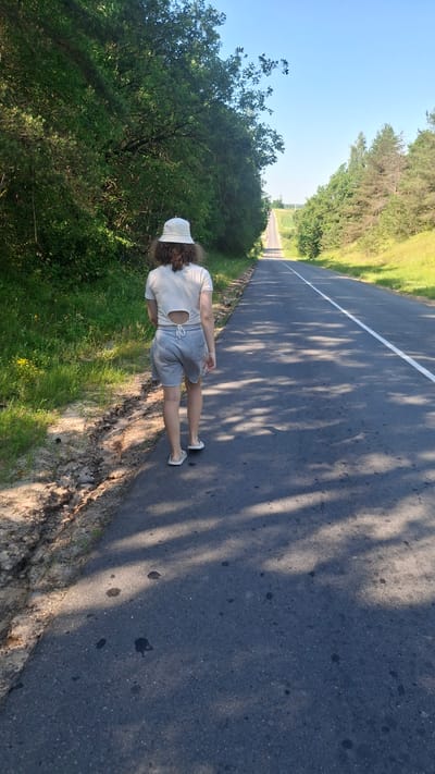A person walks away from the camera down a paved road in a rural area. The person is wearing a light-colored bucket hat, a short-sleeved t-shirt with a cutout design on the back, and grey shorts. They are also wearing white flip-flops. The road has a white line down the center and a white line on the right edge. The road is flanked by trees and grass. The sky is clear and blue. The lighting suggests it is daytime, possibly late afternoon, as indicated by the shadows on the road. The surrounding environment appears to be natural, with no visible buildings or infrastructure other than the road itself. The location is identified as Дзямідавіцкі сельскі Савет, Belarus.