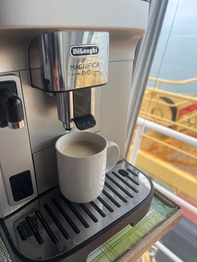 A DeLonghi Magnifica Evo automatic coffee machine, silver and black, is positioned in the foreground. A white ceramic mug containing a light brown, frothy beverage rests on the machine's black drip tray beneath the dispensing spout, from which a single drop of liquid is suspended. The machine's branding "DeLonghi MAGNIFICA EVO" is prominently displayed. To the right, a clear glass partition reveals an outdoor maritime scene under an overcast sky. A calm body of light blue-grey water extends to the horizon. Below the water line, visible structures include large, yellow-painted metallic sections with white railings and distinct red markings, showing evidence of rust and general wear, consistent with a vessel or dock facility. The depicted environment is situated in Bangor, United Kingdom, overlooking its coastal waters.