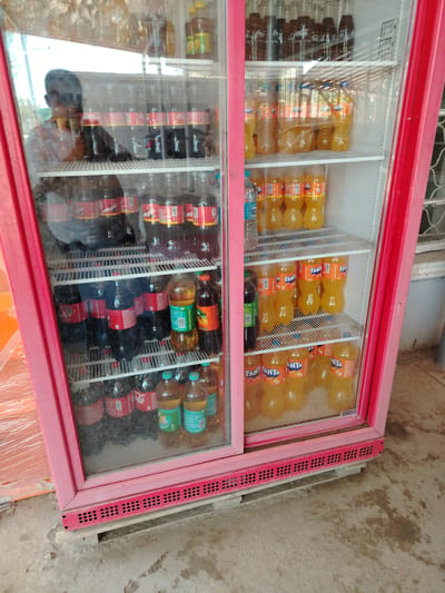 In the city of Usto Xo‘jamat, Uzbekistan, a bright pink, double-door commercial refrigerator is situated outdoors, possibly on a street or market stall, as indicated by the concrete ground and a wooden pallet beneath it. The refrigerator is filled with a variety of bottled beverages, predominantly sodas.
The left side of the refrigerator showcases rows of Coca-Cola bottles, identifiable by their red labels. These bottles are of various sizes and appear to be standard plastic bottles. There are also some darker colored bottles visible on the top shelf of this section.
The right side of the refrigerator is predominantly stocked with Fanta, recognizable by its orange color and distinct labeling. Several shelves are filled with these orange-colored Fanta bottles, also in plastic packaging. Other brands and types of beverages are visible on some shelves, including what appear to be water bottles.
The refrigerator's glass doors are partially reflective, showing a reflection of a person wearing glasses and a patterned shirt standing behind it, suggesting a vendor or shopkeeper. The interior lighting of the refrigerator illuminates the products. The overall scene depicts a commercial setting where beverages are being sold.