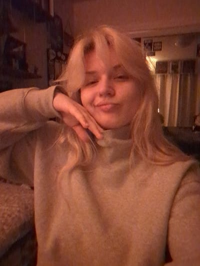 A young adult female with long blonde hair and bangs, wearing a light-colored turtleneck sweater, is depicted from the chest up in a dimly lit indoor setting. She is positioned towards the center of the frame, facing the camera, with her head slightly tilted. Her right hand is raised to her right cheek, fingers gently splayed against her skin. Her lips are pursed in a 'duck face' expression, and her eyes are open, looking forward. The background on the left is dark and indistinct, showing what appears to be a shelving unit with items. The background on the right reveals a lighter section, possibly a wall with curtains or drapes and a few framed objects. Illumination is warm-toned and appears to originate from artificial light sources. Digital noise is present throughout the image. The specific location in the city of Area D (Elaho/Garibaldi), Canada, cannot be determined from the visual information provided.