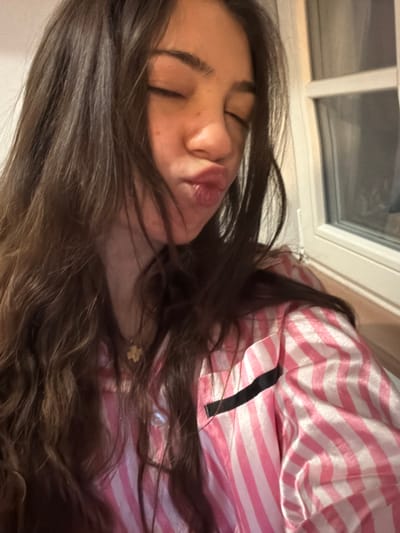 A young woman with long, dark brown hair is lying down, eyes closed, with slightly puckered lips. She is wearing pink and white striped pajamas with a black strap visible. A small gold necklace hangs around her neck. The background shows a window with a white frame. The image is rotated at an angle.