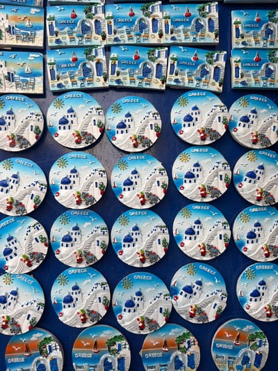 This image displays a collection of souvenir magnets, likely purchased in Keramoti, Greece. The magnets feature depictions of traditional Greek architecture, characterized by white buildings with blue domes, set against a backdrop of the sea and sky. Some magnets also include stylized representations of local flora and marine life, such as sailboats and birds. The word "GREECE" is prominently visible on many of the magnets, reinforcing their origin and theme. The arrangement suggests a retail display, possibly in a shop or market stall. The overall impression is one of a sunny, Mediterranean scene, evoking a sense of vacation and cultural tourism. The objects are vibrantly colored, with blues, whites, and touches of red and green dominating the designs. The background is a dark blue surface, likely a table or display stand.