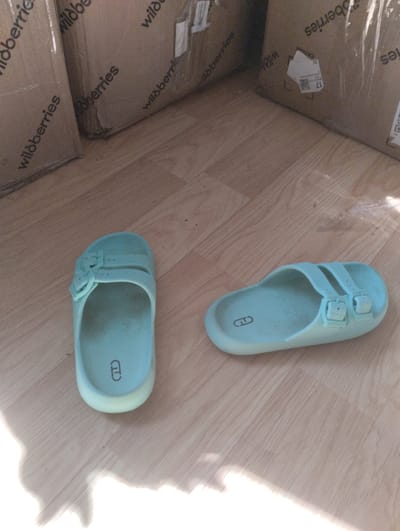 The image depicts two light blue slides resting on a wooden floor. The slides are of a simple design with two straps across the top, each secured with a buckle. A circular logo with a stylized 'G' and 'D' is visible on the insole of the slide on the left. In the background, several cardboard boxes are stacked, some of which have "wildberries" printed on them. One box in the background has a shipping label with the number "17" prominently displayed. The lighting suggests an indoor environment, possibly a room or hallway. There are no people visible in the image. The scene is static and does not show any ongoing action.