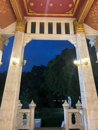 The image shows a view through an ornate gateway with ornate gold decorations on the ceiling and pillars. The sky is dark blue, suggesting it is dusk or night. Two outdoor lamps with gold bases and glass shades are visible on the pillars, casting a warm glow. Beyond the gateway, dense green trees are visible, with some grass in the foreground. The architecture appears to be of a temple or a similar cultural building. This scene is likely located in Tha Khlong Town Municipality, Thailand, given the architectural style often found in such locations.
