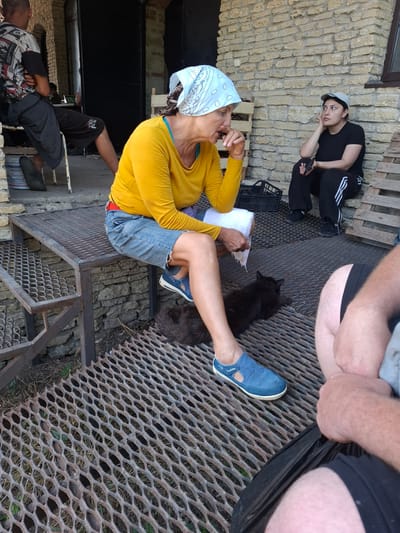 In the city of Krasnaya Skala, Russia, a group of people are gathered outdoors, near a building with a stone wall. A woman wearing a yellow long-sleeved shirt, denim shorts, and a light blue headscarf sits on a metal platform. She is holding a white cloth in her right hand and appears to be eating something. To her left, a black cat rests on the metal grating. In the background, a woman in a black t-shirt and cap sits on some steps, holding a phone. To the far left, a man in a t-shirt and shorts is seated. The environment appears to be rustic, with wooden crates visible in the background and dry grass on the ground. The lighting suggests it is daytime.