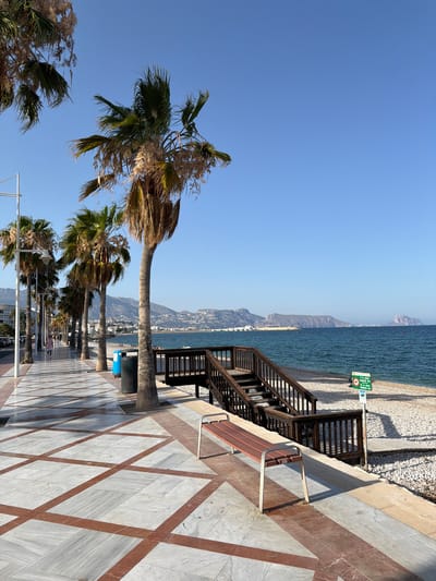 The image depicts a scenic promenade in Altea, Spain, bordering a pebble beach and the Mediterranean Sea under a clear blue sky. The promenade features a distinctive marble-patterned walkway with red-brown geometric inlays. A row of tall palm trees lines the left side of the walkway, casting shadows and enhancing the tropical ambiance. In the foreground, a wooden bench with a metal frame is positioned near the edge of the promenade, offering a view of the beach.
Adjacent to the promenade, a wooden staircase leads down to the pebble beach. The beach itself is wide and extends to the sea, which appears calm and deep blue. In the background, the coastline of Altea is visible, characterized by buildings nestled against rolling hills and mountains. The distant mountains, bathed in sunlight, form a striking backdrop to the coastal scene.
Several objects are strategically placed along the promenade. Blue trash receptacles are visible near the palm trees, suggesting maintenance and public service. A green sign with pictograms indicating rules for the beach is posted near the wooden stairs. The water is a vibrant blue, and the sky is an unblemished expanse of clear blue. The overall impression is one of a serene and well-maintained coastal area, popular for leisure and relaxation.