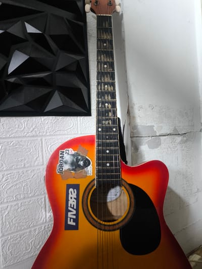 The image shows an acoustic guitar leaning against a white textured wall, possibly in the city of General Trias, Philippines, based on the context provided. The guitar has a sunburst finish, transitioning from red to orange to yellow, and features stickers on its body. One sticker prominently displays "MICHAEL JORDAN 23 GREAT" with a black and white image of the basketball player. Another sticker is blue and has white lettering that appears to read "FIVB92" in reverse. Behind the guitar, to the left, is a black, geometric art piece mounted on a white brick-patterned wall. To the right of the guitar, the white wall shows signs of wear and peeling paint, with a visible crack. The lighting suggests an indoor setting. There are no people visible in the image.