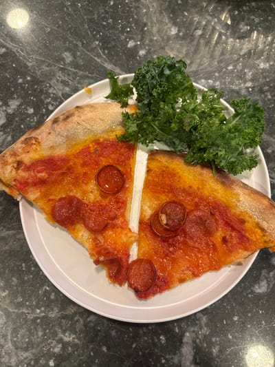 The image displays two triangular slices of pepperoni pizza alongside a pile of green kale, arranged on a white, circular plate. The pizza slices feature a golden-brown crust, a red tomato sauce base, orange-yellow melted cheese, and several reddish-brown pepperoni discs, some exhibiting curled edges and rendered fat. The dark green, curly kale leaves are situated at the top of the plate, partially overlapping the pizza crusts. The plate rests on a dark, mottled countertop, consistent with granite or quartz, which shows a reflective sheen from an overhead light source in the upper left. No individuals or dynamic actions are present. The exact location within Rolling Hills Estates, United States, cannot be determined from the visual data provided.