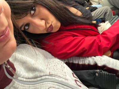 Two women pose closely for a selfie. One is partially visible, only her lips are in frame. The other gazes at the camera with dark eyes and pursed lips. She wears dark hair with bangs and a red jacket. They appear seated, likely on a mode of transportation, surrounded by other people and luggage. The first woman wears a gray jacket with a visible zipper.