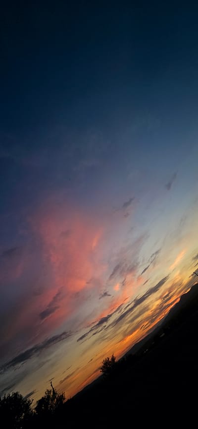 The image depicts a vibrant sunset over a rural landscape, likely in or near Momin Prohod, Bulgaria. The sky is filled with dramatic clouds, transitioning from a deep navy blue at the top to hues of pink, orange, and yellow near the horizon. Wispy clouds are scattered throughout, some catching the sunset's light and appearing as streaks of color. In the foreground, silhouetted trees and a darkened landscape suggest a rural or mountainous terrain. The perspective is tilted, giving a dynamic feel to the scene. No people or specific structures are clearly identifiable, focusing the image's attention on the natural beauty of the sunset.