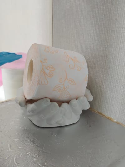 An overhead shot of a toilet paper roll with an orange floral pattern is resting on a white ceramic holder shaped like hands, situated on a textured surface. In the background, out of focus, are other rolls of toilet paper, one pink and one blue. The scene appears to be an indoor setting, possibly a bathroom or storage area. There are no people or animals present. The lighting is soft and even, casting no harsh shadows. The image does not provide any information that would allow for its localization to Dzerzhinsk, Russia.