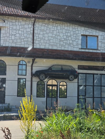 The image depicts a building with a dark-colored car mounted on its facade. The building features a white exterior with a geometric pattern on the upper section and a light-colored plaster on the lower section. Several windows of varying shapes and sizes are visible, including arched windows and rectangular ones with dark frames. The car is positioned horizontally on the wall, appearing to be a hatchback. In the foreground, there is lush green vegetation, including a tall plant with yellow flowers and some grass. The scene appears to be an outdoor setting, possibly in the city of Bregovo, Bulgaria, given the context. There are no people visible in the image.