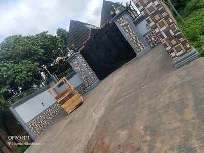 The image shows a large, ornate black metal gate, flanked by decorative pillars. The pillars are made of grey concrete and adorned with a pattern of alternating brown, beige, and grey square tiles. Above the gate, a dark, angled roof with dark grey tiles is visible, with smaller decorative elements and lighting fixtures. To the left of the gate, a grey wall with a similar tiled pattern runs along the property line, topped with razor wire for security. Lush green trees and vegetation are visible behind the wall and to the right of the pillars.
In front of the wall, to the left of the gate, rests a wooden crate. The ground in the foreground is a worn, greyish-brown concrete surface, showing signs of wear and tear with scuff marks and some dirt. The sky above is overcast, with a mix of white and grey clouds, suggesting a cloudy day.
The overall impression is of a secure and well-maintained property entrance in a semi-tropical or rural environment. The location is identified as "Nigeria, Obodoukwu" and the camera information "OPPO R11" is visible in the bottom left corner. No people or specific activities are depicted.