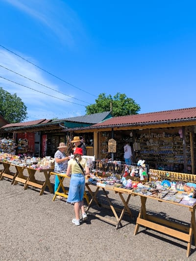 The image depicts an outdoor market scene in Khotyn, Ukraine, under a clear blue sky with faint wispy clouds. A row of low-rise market stalls, featuring wooden structures and corrugated metal roofs in reddish-brown and dark grey, stretches across the midground. In front of these stalls, multiple wooden trestle tables are set up on a light-colored paved or gravel ground, covered with a wide array of merchandise including keychains, small ceramic items, decorative plates, plush toys, and various tourist souvenirs.

Three individuals are prominently visible in the central area. On the left, a female vendor, wearing a patterned top and a straw hat, is interacting with a female customer, seen from behind. The customer is dressed in a yellow t-shirt, blue denim jorts, white sneakers, and a red bucket hat, and is observed examining items on a table. Behind them, a male in a dark shirt and straw hat stands near the stall entrance. Further to the right, inside one of the stalls, another person in a white top is partially visible amidst shelves filled with goods. Green deciduous trees are present in the background above the stalls, and several dark utility lines are strung across the upper portion of the sky.