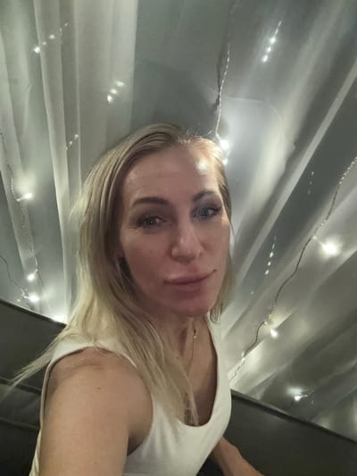 A woman with blonde hair and blue eyes is pictured in a selfie, wearing a white tank top. She appears to be indoors, with sheer white curtains and string lights in the background. The lighting suggests a dim, possibly evening setting. There is no information to suggest this image is located in Starye Chernitsy, Russia.