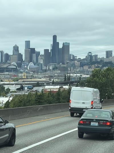 The image shows a view of the Seattle skyline from a highway, likely during the daytime under overcast skies. In the foreground, several vehicles are visible on the road, including a white van and a dark-colored sedan. The sedan has a license plate that reads "LM8852". The highway appears to be elevated, with a concrete barrier separating the lanes from the embankment and trees to the left. Beyond the highway, the dense urban landscape of Seattle unfolds, characterized by numerous tall buildings and skyscrapers of varying architectural styles. The city appears to be situated by the water, though the water itself is not clearly visible. The overall atmosphere is that of a typical commute on a cloudy day in a major metropolitan area. The image is oriented vertically, suggesting it was taken while the photographer was looking out a window.