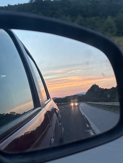 The image shows a view from a car's side mirror, capturing a sunset scene on a highway. In the foreground, the reflection of the car's dark red body is visible, along with parts of its window and frame. The mirror reflects the road ahead, where another vehicle with illuminated headlights is driving. The sky exhibits a warm gradient of colors, transitioning from orange and pink hues near the horizon to lighter blues and grays higher up, indicative of dusk. Trees and hills form the background on either side of the highway. The overall environment appears to be a rural or semi-rural setting, possibly on the outskirts of a town or city. The specific location in Kostadinkino, Bulgaria is not visually discernible from the image itself.
