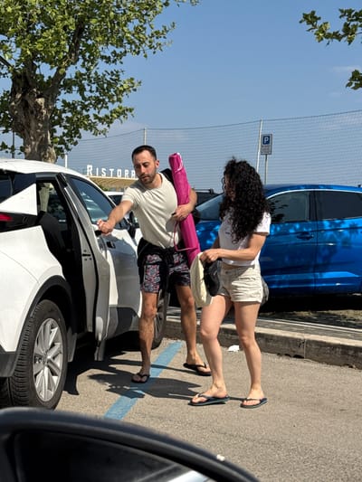A couple is interacting next to a white car in a parking lot in Anzio, Italy. The man, wearing a white t-shirt and patterned shorts, is opening the car door, holding a rolled-up pink item (possibly a beach umbrella) under his arm. The woman, with curly black hair, wears a white top and light-colored shorts; she appears to be placing items (a black shoe and a beige item) into a bag. Both are wearing flip-flops. A blue car is parked behind them, and a tree is on the left side of the image. A partially visible parking sign with a "P" is also visible. The word "RISTORA" is partially seen in the background. Another car is in the foreground.
