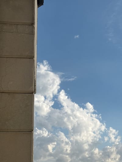 This is a vertical, medium shot photograph taken from below, looking up at the sky from a city street in Abovyan, Armenia. The left side of the frame is dominated by a cream-colored stone building or structure, composed of at least three stacked rectangular blocks. The stone appears aged and shows some signs of cracking. The top edge of the frame reveals a dark, sloped element, possibly part of a roof or overhang.

The right two-thirds of the image is a clear blue sky filled with white, fluffy cumulus clouds. The clouds vary in density, with some appearing wispy and others more substantial. A small, isolated cloud is visible higher up in the sky, to the left of center. The sunlight suggests it is daytime, and the overall impression is one of an outdoor urban environment. No people or animals are visible in the frame.