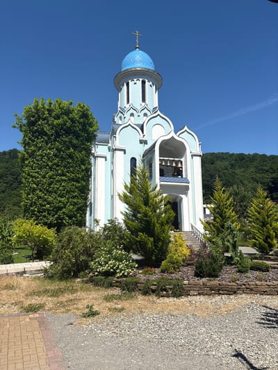 The image shows a light blue and white church with a blue dome, topped with a cross. The church has a bell tower with multiple bells. It is surrounded by lush green trees and manicured landscaping, including a stone wall and gravel paths. In the background, a densely forested hillside rises under a clear blue sky. The scene appears to be peaceful and well-maintained, suggesting it is a place of worship located in a natural setting. The location is identified as the city of Lesnoye, Russia. There are no people visible in the image.