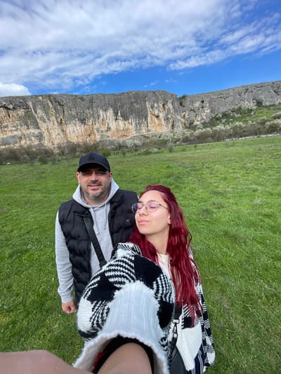 The image depicts a man and a woman lying on a grassy area near a cliff. The man is wearing a black cap, a gray hoodie, and a black vest. The woman has long, red hair and is wearing glasses. They appear to be posing for a selfie. A black and white sweater is on top of the man. In the background, there is a rocky cliff face under a blue sky with some white clouds. The photo was probably taken in the city of Barkach, Bulgaria, where similar cliffs exist.