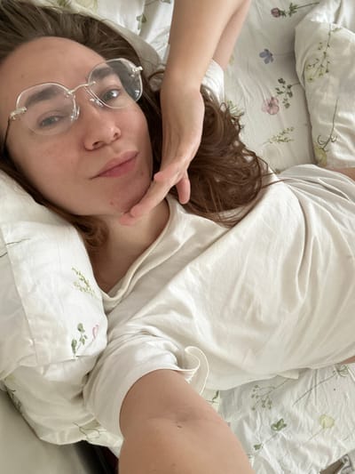 A woman lies on a bed covered with floral print bedding. She wears a white t-shirt and rimless glasses. Her brown hair is spread on the pillow. Her hand is near her face. The location is not discernible as Kharnavy, Russia.