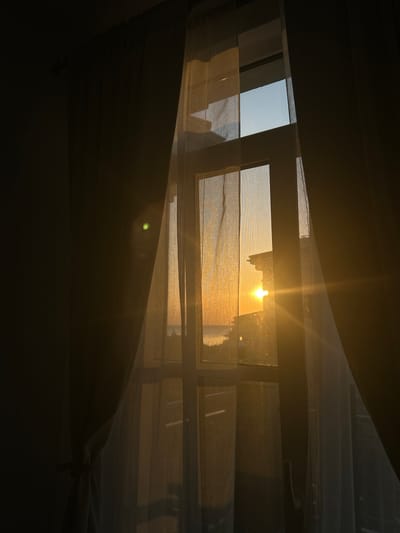 The image depicts a window overlooking a sunset, with curtains framing the view. The sun is setting over the sea, casting a warm, golden light that is filtered through the sheer curtains. A building is visible in the distance, silhouetted against the bright sky. The scene is peaceful and serene, evoking a sense of calm and tranquility. The location is the city of Chernomorets, Bulgaria, a coastal town known for its beautiful beaches and sunsets.