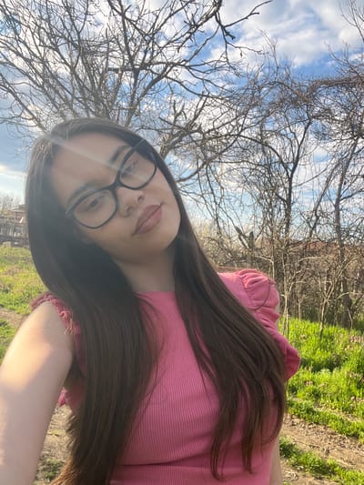A selfie portrait of a young woman is presented at an angle. The woman is wearing glasses and a pink top. She has long, dark hair. In the background, there are trees without leaves, a blue sky with clouds, and some green grass. There is no indication of Blaskovo, Bulgaria in the image.
