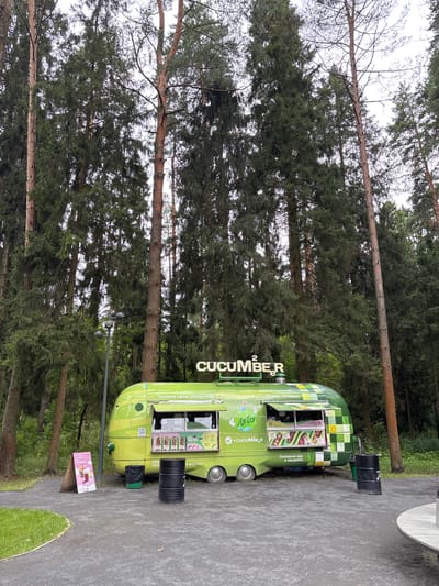 A bright green food truck branded "CUCUMBER" is parked on a paved path in a forest clearing. The food truck is adorned with colorful graphics and has an open window displaying images of food items and pricing. Two black cylindrical trash cans and one green trash can are placed next to the food truck. A triangular A-frame sign with graphics of drinks and berries stands to the left of the food truck. Tall, mature pine trees with dense foliage surround the clearing, indicating an outdoor, possibly park-like, setting. The ground is covered with a dark gray textured surface, transitioning to green grass on the left side of the frame. A partial view of a light-colored round table with a silver leg is visible in the lower right corner. The sky appears overcast. The location is identified as Pavlovskaya Sloboda, Russia. There are no people visible in the scene.