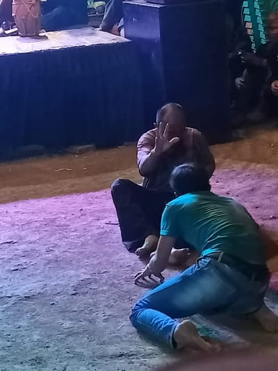 This image captures a dramatic scene, likely a performance or ritual, taking place at night in Kecamatan Cimaung, Indonesia. In the foreground, two men are positioned on what appears to be a dirt or concrete ground, illuminated by artificial blue and purple lighting.

An older man, bald and wearing a dark t-shirt and dark pants, sits cross-legged, facing partially towards the viewer. His right hand is raised, palm outward, in a gesture that could convey stopping, defense, or surprise. His expression is partially obscured but suggests intensity.

In front of him, a younger man, wearing a green t-shirt and blue jeans, is on his hands and knees, facing away from the viewer and towards the older man. His posture suggests crawling or reaching, possibly in supplication or an aggressive movement, making for a dynamic interaction between the two figures. Both men are barefoot.

In the background, on the left, a raised platform or stage is visible, draped with dark fabric. A light-colored, drum-like instrument sits on the platform. Further back and to the right, other indistinct figures and dark structures, possibly sound equipment or parts of the stage setup, are visible, partially obscured by the darkness of the night. The overall impression is one of a theatrical or ceremonial event under artificial illumination. No visible text is present in the image.