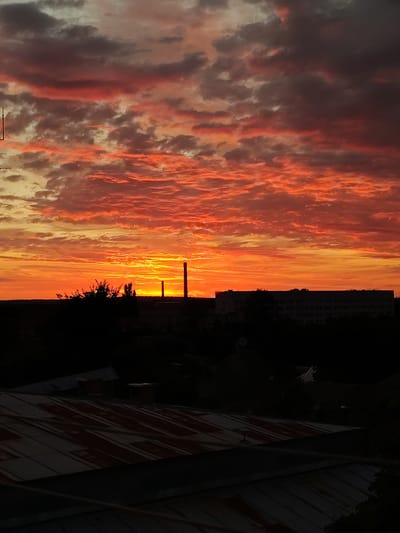 The image captures a dramatic sunset over the city of Okhtyrka, Ukraine. The sky is ablaze with vibrant hues of orange, red, and yellow, with clouds casting a dramatic shadow across the landscape. Silhouetted against this fiery backdrop are industrial structures, including two tall smokestacks, and a long, rectangular building, possibly a factory or administrative complex. A cluster of trees and other buildings are visible in the foreground, also in silhouette, suggesting the image was taken from an elevated vantage point. The bottom of the frame shows the roofs of several buildings, some appearing to be aged or dilapidated. There are no people or animals visible in the image. The scene is devoid of any activity or interaction, presenting a static, atmospheric view of the city at dusk.
