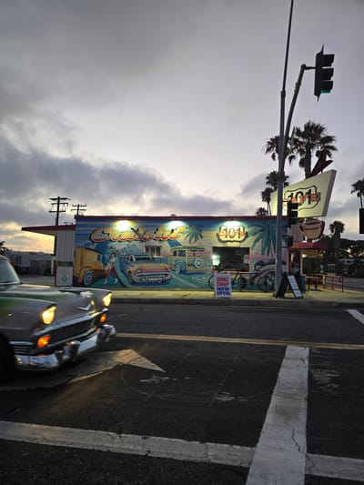 The image shows a street scene in Oceanside, California, at dusk. A classic car is in the foreground, driving on a road. In the background, there is a building with a mural depicting surf culture and vintage cars. The building appears to be a cafe called "101 Cafe". There are palm trees and a traffic light visible, with the green light illuminated. A sign on the sidewalk reads "SMOKE SHOP NOW OPEN". The sky is overcast, with hints of yellow from the setting sun.