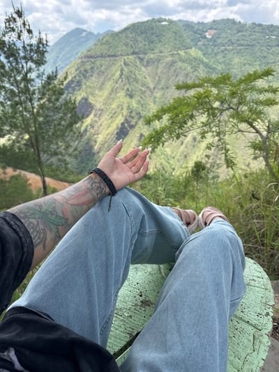 A first-person perspective shot shows a person seated outdoors in Lũng Cú Commune, Vietnam. The person wears light blue jeans and sandals, with an arm resting on the leg, revealing tattoos and a bracelet. A light green, weathered wooden seat is visible. The background features a mountainous landscape with lush green vegetation under a cloudy sky.