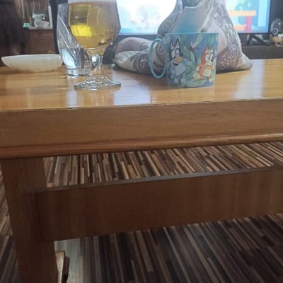 The image shows a wooden table with a glass of beer, a white dish, a blue mug with cartoon characters, and a patterned pillow. The table appears to be in a home environment, possibly a living room or dining area, with a television visible in the background. The floor beneath the table has a striped pattern. There is no indication of the city of Lyaskovets, Bulgaria.