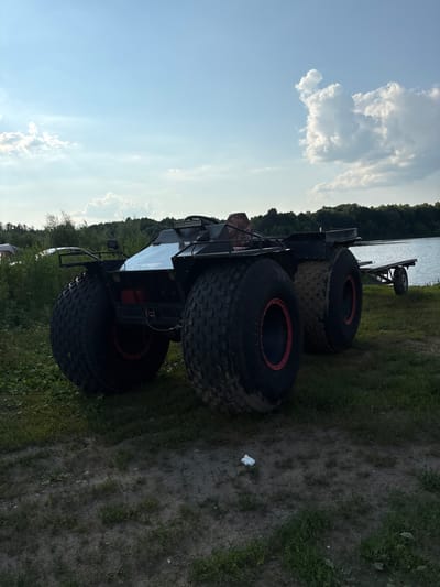 This media file, taken in Gur'evo (Volkovskii S/S), Russia, shows an all-terrain vehicle (ATV) parked on a grassy embankment beside a body of water. The ATV is large, black, and equipped with exceptionally wide, low-pressure tires with red rims, suggesting it is designed for challenging terrain. It has a rack on the front and what appears to be a roll cage. A small trailer with a single wheel is hitched to the rear of the ATV.
The scene is set outdoors, with the water appearing to be a lake or river, bordered by a line of trees in the background. The sky is a clear blue with some scattered white clouds, indicating fair weather. The lighting suggests it is daytime, likely late afternoon or early evening due to the angle of the light and shadows. The ground is a mixture of grass and dirt, with some areas of mud or wet soil around the ATV's tires. There is a small piece of white debris on the ground near the ATV. No people are visible in the frame. The overall impression is one of recreational use or readiness for off-road exploration.