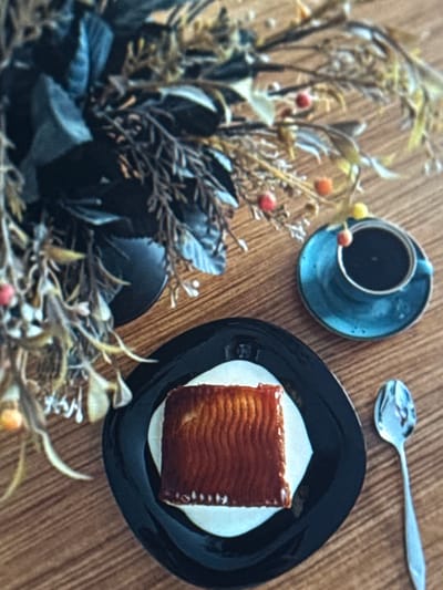 This image shows a close-up overhead view of a dessert and coffee on a wooden table. The dessert is a rectangular piece of cake with a layered, wavy texture on top, placed on a white paper doily on a black plate. A silver spoon rests above the plate, angled towards the left. To the left of the dessert, a blue ceramic coffee cup sits on a matching saucer, filled with dark liquid. The coffee cup is adorned with small red and yellow berries. A decorative arrangement of dried plants and dark foliage occupies the left side of the frame, adding a natural element to the scene. The wooden surface of the table has a visible grain pattern. The lighting suggests a natural, possibly indoor, setting. No people or visible text are present in the image.