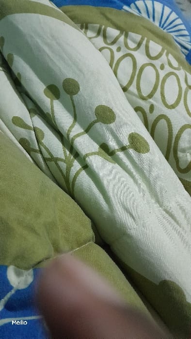 The image shows a close-up of a patterned bed sheet in green and white, with a blue bed sheet partially visible in the bottom left corner. The sheet features a repeating pattern of stylized green flowers. The bed sheet is folded and the photo was taken from a low angle, capturing part of an arm in the lower right corner. The image was likely captured inside a home located in Kapsabet, Kenya, based on the provided coordinates.
