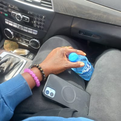 In the city of Dorowa, Nigeria, a close-up shot reveals a person's hand holding a blue plastic bottle of Swan water. The hand, adorned with a gold ring on the index finger and two beaded bracelets (one pink and one black with gold accents), is positioned over a black phone with a textured case, which is resting on the person's lap. The background includes the interior of a car, with a portion of the dashboard and gear shift visible. The overall scene captures a moment of casualness, possibly during a car ride.