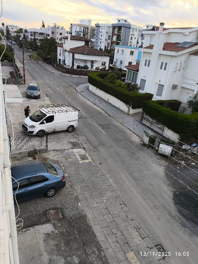 An elevated, downward-angled color photograph captured on 13/11/2025 at 16:05 depicts a residential street scene in Nicosia, Northern Cyprus / Cyprus. The asphalt road curves gently to the right in the mid-ground, flanked by paved sidewalks.
On the left side of the road, a white commercial van with a black roof rack is parked, facing away from the camera. One individual, dressed in dark clothing with a visible reflective element, stands next to the driver's side of the van, facing away from the camera. Further up the street, a dark grey sedan is parked behind the white van. In the lower left foreground, a dark blue sedan is partially visible, parked on a paved area adjacent to the viewpoint structure. Utility poles and overhead wires run along the left side of the street.
On the right side of the street, two large green refuse bins are positioned near a pile of assorted refuse. The sidewalk is bordered by a well-maintained green hedge atop a white wall. Behind the hedge, a prominent white two-story house with a red-tiled roof and solar water heating panels is visible, along with a metal gate. In the background, a mix of multi-story modern apartment buildings, predominantly white with grey and brown accents, and additional single-story houses with tiled roofs, populate the urban landscape. The sky above is partially cloudy, showing hues of light blue and soft orange, indicative of late afternoon lighting. The scene appears generally orderly, save for the refuse pile.