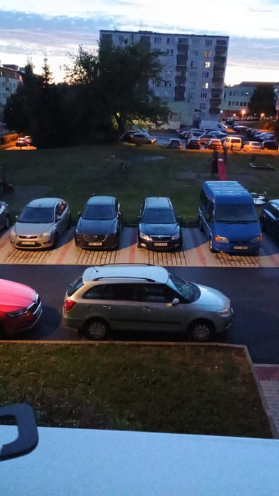 The image shows an outdoor scene at dusk, likely in Sokolov, Czechia, as indicated by the presence of a residential apartment building typical of Czech architecture and cars with Czech license plates. In the foreground, a silver station wagon is parked. Behind it, a row of parked cars includes a silver hatchback on the left, a dark hatchback in the middle, and a black hatchback on the right. Further back, a blue van is parked. The background features a large apartment building with illuminated windows, trees, and a sky with clouds. A playground slide is visible in the middle ground. The overall scene depicts a residential area at the end of the day.