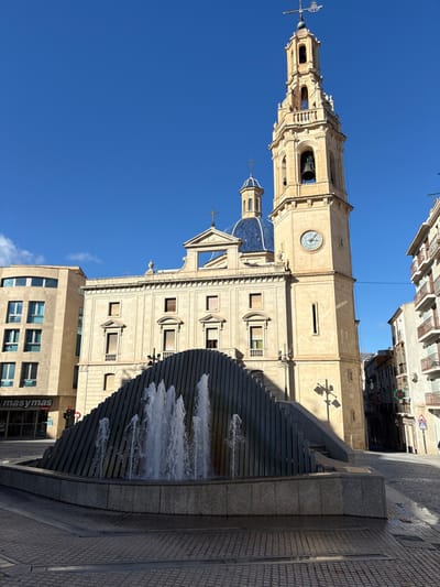 The image displays a clear, sunny urban scene in Alcoy/Alcoi, Spain, centered on a public square. In the immediate foreground, a multi-tiered, dark gray angular fountain with vertical slats actively sprays multiple jets of water into a stone basin. Behind the fountain, the prominent Co-cathedral of Santa Maria features a light-colored stone facade, a tall, ornate bell tower with a visible clock face, a large bell, and a weather vane on its spire. A distinct blue-tiled dome with a cross is visible behind the main church structure.
To the left of the church, a modern, light-colored building houses a "masymas" store on its ground level. To the right of the church, an older street descends, flanked by multi-story residential or commercial buildings with balconies. The ground is paved with patterned pavers and cobblestones. Long shadows are cast from the left, indicating bright sunlight. A red traffic light and a street lamp are visible elements of the urban infrastructure. No individuals are present in the scene.