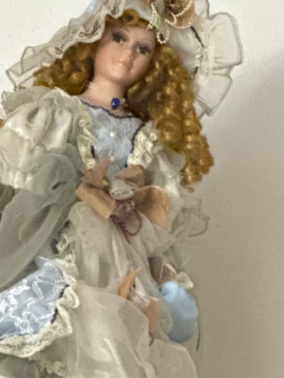 The media file features a close-up, overhead shot of a porcelain doll lying amidst a collection of white and pastel-colored fabrics. The doll has a porcelain face with painted features, including dark eyes and rosy cheeks, framed by curly, golden-brown hair. She wears an ornate dress with lace trim and appears to be adorned with a blue jewel pendant around her neck. The doll is surrounded by layers of fabric that resemble doll clothing or bedding, with ruffles and sheer material creating a soft, elaborate presentation. Some of the fabrics are a pale blue, adding to the pastel color scheme. There are small, light-colored decorative elements scattered around, possibly ribbons or small fabric flowers. The background is a plain, light-colored surface, likely a wall or table, with a slight texture. The overall impression is of a static display, possibly a doll laid out for viewing or as part of a decorative arrangement. There are no discernible human figures actively engaged with the doll. The lighting is soft and diffused, suggesting an indoor setting. No text is visible in the image. The scene's context is Borgosatollo, Italy, but this information does not offer specific visual cues within the image itself.