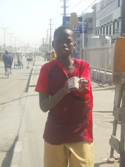 A young male stands centered on a concrete sidewalk in the city of Babban Giji, Nigeria, facing the viewer directly. He has dark skin, short dark hair, and wears a two-toned short-sleeved shirt, red on the upper chest and shoulders, and maroon on the lower torso. The shirt appears worn, with visible fraying. He also wears light tan shorts. Both hands are held forward, presenting a small stack of folded banknotes, with the number "500" partially visible on the top note.

To the male's immediate right, a wooden support structure for an orange cylindrical object is partially visible. Behind him, a wide, dusty asphalt road extends into the distance under a bright, hazy sky. On the opposite side of the road, a multi-story commercial building features the text "HYPERMARKET" and "24 OPEN" on its facade. Adjacent to this building, a blue street sign displaying "RUHODA YOGURT SARA" is visible, with a light-colored cardboard cutout attached above it. Overhead power lines and multiple utility poles traverse the upper portion of the scene.

On the road, a person on a bicycle, wearing a blue top, travels away from the camera on the left side of the street. Further down the street, various pedestrians and vehicles, including several motorized tricycles, are observed. Numerous streetlights with multiple fixtures line both sides of the road, which appears to lead towards a distant pedestrian bridge or overpass. The environment suggests a busy urban commercial area.